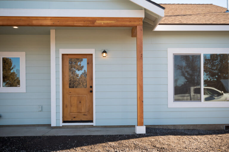 Charming facade of a modern ADU with light blue siding, white trim, large windows reflecting trees, and a covered entry with exposed wood beams.