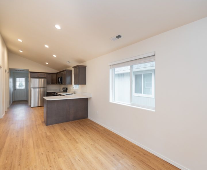 Minimalist living area with light wood floors, white walls, and recessed lights, opening to a compact kitchen with island and stainless appliances—ready for customization.